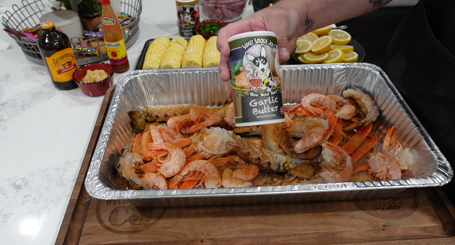 A hand holds a shaker of Sweet Smoked Joy Garlic Butter seasoning over an aluminum pan filled with cooked lobster tails and shrimp.