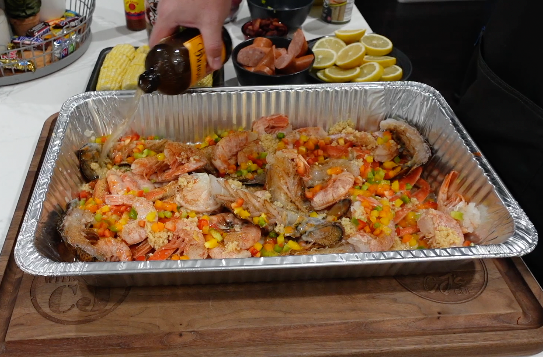 A hand pours liquid from a bottle into an aluminum pan filled with seasoned shrimp, diced bell peppers, and garlic.