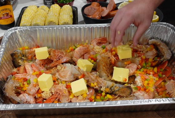 A hand places cubes of butter onto a large tray of raw shrimp and diced bell peppers, with corn and sausage in the background.