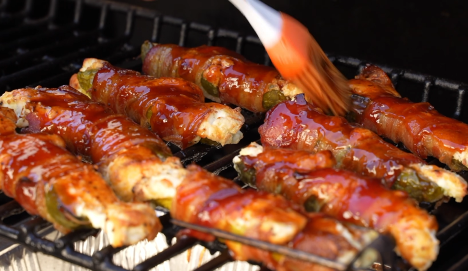 A basting brush coats bacon-wrapped jalapeño poppers with barbecue sauce on a grill.