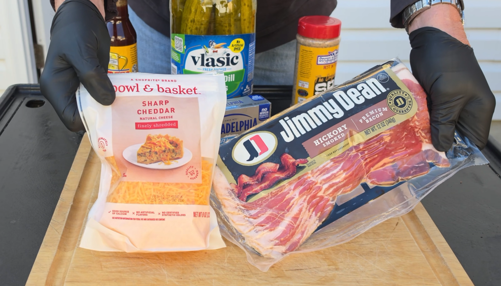 Hands in black gloves hold a bag of shredded sharp cheddar cheese and a package of hickory smoked bacon over a wooden cutting board with a jar of pickles and spices in the background.
