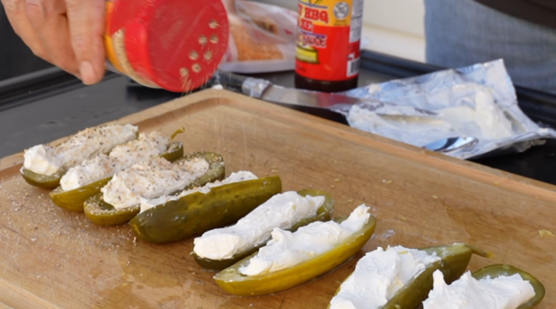 A hand sprinkles seasoning over several pickle halves filled with cream cheese on a wooden cutting board.