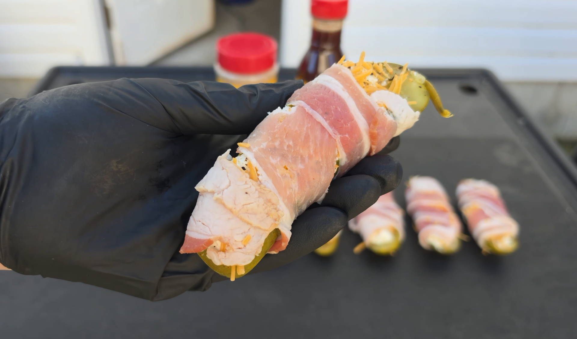 A gloved hand holds a pickle wrapped in turkey and bacon, with more prepared rolls on a grill in the background.
