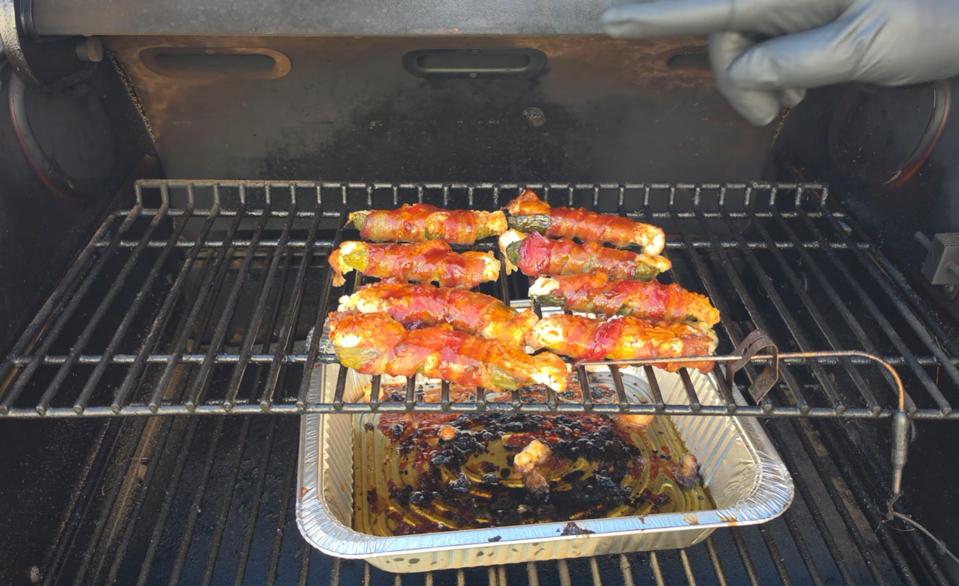 Bacon-wrapped jalapeño poppers cooking on a grill rack above a foil drip pan.